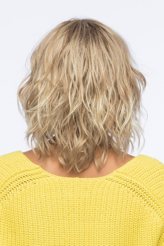 Back view of woman modeling a dark blonde wig with light blonde highlights and golden brown roots.